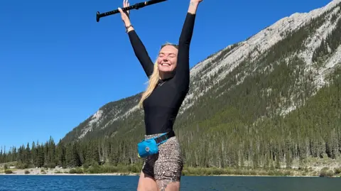 Georgie Swallow Georgie on a paddleboard on a lake, holding her paddleboard in the air