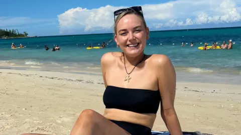 Georgie Swallow Georgie in a black bikini on a beach, smiling at the camera with the sea behind her