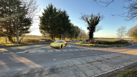 Google A street view image of the junction which forks off to the left. A brick wall runs along the side of the road to the right with tall trees behind it and trees and grass on the left-hand side of the road.