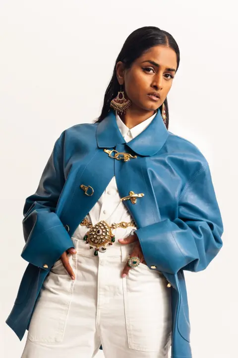 Megan Winstone Priya Ragu dressed all in white with a blue overcoat. Her long dark hair is tucked behind her ears and she is wearing bold, gold earrings