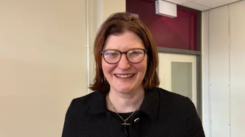 A woman with shoulder length light brown hair smiles toward the camera. She is wearing dark rimmed glasses, a black top and a black blazer.