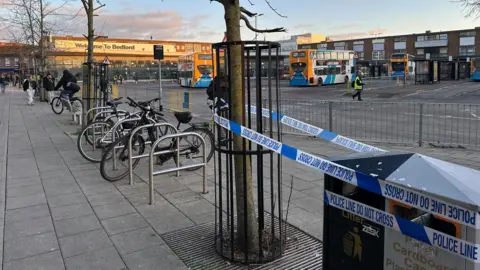 Kimberley Piper/BBC Police tape that says "POLICE LINE DO NOT CROSS" tied round a tree and a bin near Bedford bus station. There is a bike rack next to the cordoned-off tree. Buses are parked up at the bus station in the background
