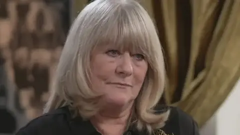 Christine Flack looks to the right of the camera as she is being interviewed. She has a solemn expression and is wearing a dark blouse. She has long blonde hair with a fringe. 