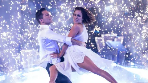 Guy Levy/BBC Pasha Kovalev, in a white shirt and dark trousers and Caroline Flack, in a white floaty dress, in mid-dance, with Caroline lifted by Pasha as sparkling white lights form a backdrop