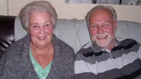 Family handout Mary with husband Ron. Ron died in 2016. They are both sat on a sofa smiling.