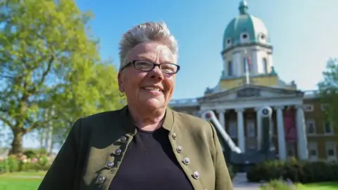 Pte Carol Morgan stands in front of the Imperial War Museum, smiling slightly off camera. She's wearing an olive green jacket and black t-shirt, and has short blonde hair and glasses.