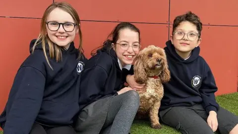 Eleven-year-old Betsy, 10-year-old Kayla, Poppy the dog, and 11-year-old Henry sit on a block covered with artificial grass. Kayla is leaning towards Poppy and Henry has an arm around the dog's neck.