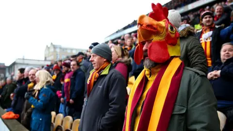 Getty Images Bradford City fans stand up in the stands to watch a football match, many wear red and yellow club scarves and a man at the front wears a bantam mask