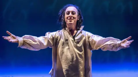 PA Media Alan Cumming on stage in Burn in 2022 - he has his arms out in a gesture that suggests he's welcoming people, while dressed in a somewhat tatty looking white outfit.