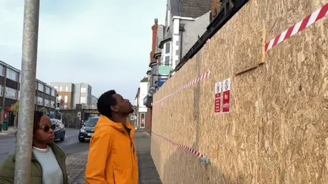 George Carden/BBC Simba wearing a yellow rain coat looking pensively up at the remains of his burnt out flat. There is a large plywood wall erected around the shop which is on the ground floor and there are charred bits of wood above the wall
