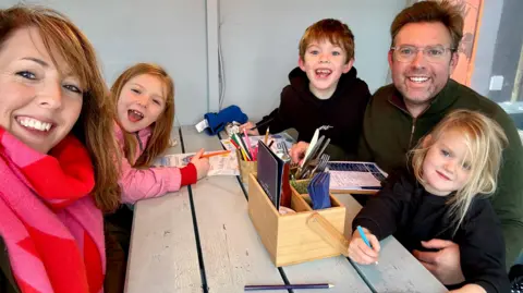 Katie and Tom The family sit around a table in a restaurant. Mum Katie and dad Tom are with their three children, a boy and two girls.