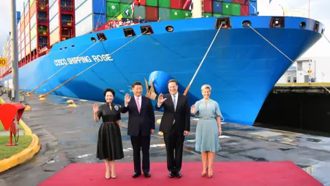 Getty Images China's President Xi Jinping and Panama's Juan Carlos Varela, wearing dark suits and ties, stand between the nations' First Ladies in formal dresses, in front of a big blue Chinese container vessel at the Cocoli locks in the Panama Canal, on 3 December 2018.