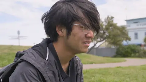 Junjie, wearing a black top and black windcheater, sits on a grassy field and cries. His hair is longer than in the first photo and he is wearing glasses.