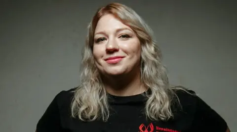Underground Standup Hanna Kochehura, a blonde lady wearing winged eyeliner, smiles at the camera wearing a black t-shirt.