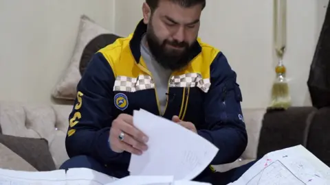 BBC News Man wearing White Helmets uniform - navy blue and yellow - looking through documents and maps.