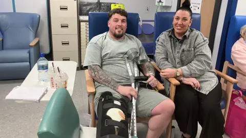 Nathan Davies has short brown hair and is wearing a grey t-shirt and shorts. He is in a chair holding crutches and has his leg strapped up. His partner is sitting next to him and has dark hair in a bun and is wearing a grey denim shirt and black trousers.