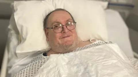 Simon Morris has grey hair and glasses and is wearing a white hospital gown and is lying in a hospital bed.