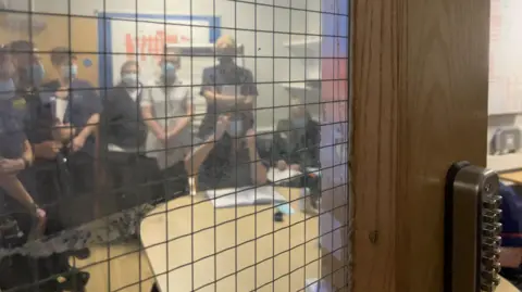 A staff meeting seen through a window in a fre doot. Staff in uniform wearing PPE stand around a desk.