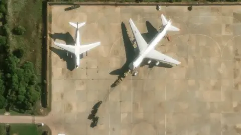 Maxar Two planes are parked on the runway at Hmeimim air force base. Military vehicles can be seen driving onto one of the planes, with further vehicles parked behind.