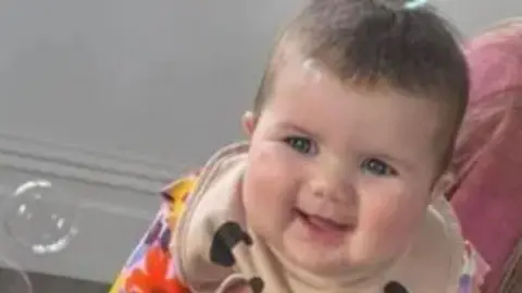 Family photo Mabli Hall pictured smiling. She is wearing a beige bib and an orange and pink and yellow top underneath