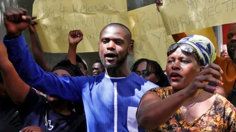 Reuters A of group of Kenyan protesters holding yellow hand-written posters aloft calling for the release of people who have been abducted. Several - including a young bearded man in a blue collars shirt and a woman wearing a brown patterned V-neck dress, necklace, blue-and-white headscarf and sunglasses on her forehead in close-up - have their fists raised.