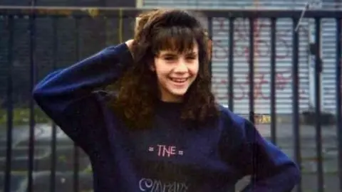 A photo of Caroline Glachan. She is a teenager posing outside in front of an iron railing. Behind the rail is a set of iron shutters covered in graffiti. Caroline is wearing a blue sweater and smiling at the camera. She has one hand behind her head.