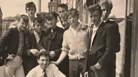 Stewart Wright A black and white photo of lots of young men gathered round a squatting man in a white shirt and black tie in a town centre.