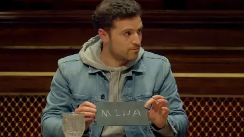 BBC/Studio Lambert Alexander holding blackboard with Mina written on it