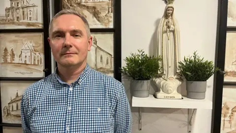 John Murphy A man with short grey hair in a white and blue checked shirt stands in front of a series of paintings of churches, a statue and some potted plants