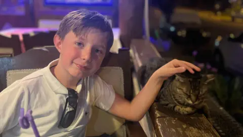Handout A young, smiling boy sits in a chair with sunglasses tucked into his white T-shirt. He has a hand on a tabby cat that is resting on a brown ledge. A purple straw is in the foreground.