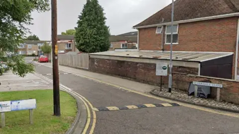 Google Silverdale Close, which has yellow and black speed humps and small blocks of flats .There is a tall conifer tree and a red car in the distance.