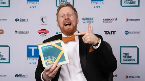 Alex Brenner Michael Campbell is wearing a black suit, white shirt and orange bow tie. 
He is seen holding the Judges' Award from the Stage Theatre Awards.