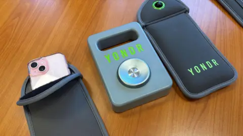 BBC Two grey neoprene phone case sits on a brown wooden table. A phone with a pink case can be seen inside one. A metal magnet remover sits in between. It has the word 'YONDR' printed on the front in green text.