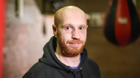 BBC Billy Ramsay looks into the camera while standing in a boxing club - a punch bag can be seen blurred in the background. Billy has a bald head, a ginger beard and moustache and is wearing a black hoodie.