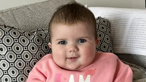 Family photo Mabli Hall sitting propped up in a grey sofa with black and grey patterned cushion. She is wearing a pink jumper