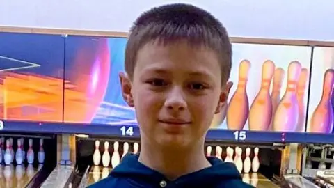 Family Leo looking straight at the camera. Ten-pin bowling is behind him with an alley leading to a set of pins.