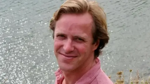 Lady Gabriella Kingston Thomas Kingston - a man in his 40s with dark blond hair - smiles as he stands near a lake. He is wearing a pink cotton shirt.