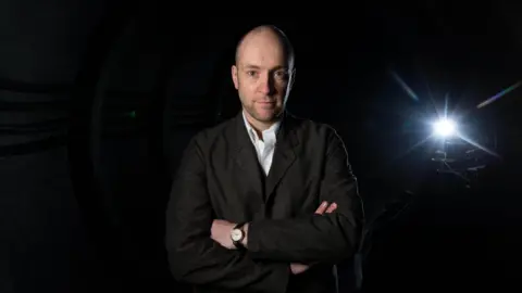 PA Media Derren Brown. He is pictured with his arms folded, looking straight at the camera with a slight smile. He is bald and has some stubble on his chin. He is wearing a dark grey jacket, a white shirt and a watch.