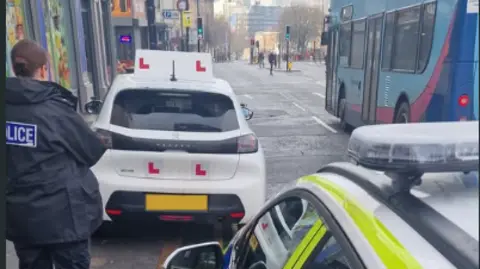 Merseyside Police Police officer taking notes on the parking of the white learner car covered with L plates, which is parked partly on the pavement over double yellow lines