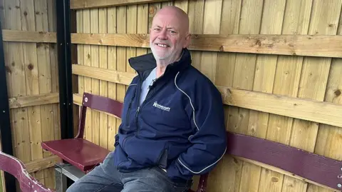 Paul O'Gorman/BBC Ian Nutter sits in the timber clad stand. He is sitting on a burgundy seat and it wearing jeans and a blue zip up casual jacket. He is bald with a short white beard and is smiling.