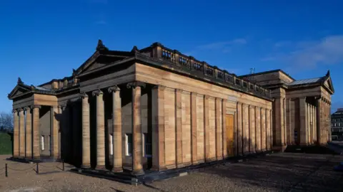 Getty Images A general view of the National Gallery of Scotland in Edinburgh. It is a sunny day with blue sky and a few grey-white clouds in the sky. The building itself is made from light stone and has two sets of four columns on either side.