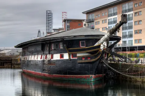 Unicorn Preservation Society A large black ship with white and red detailing ship dating from the 1800s sits in a dock, with a modern apartment building in the background