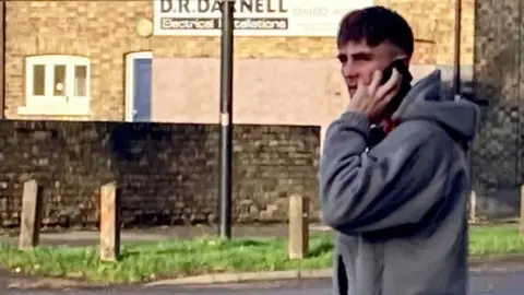 Brian Farmer/BBC A man wearing a grey hooded top holding a mobile phone to his left ear with his left hand.