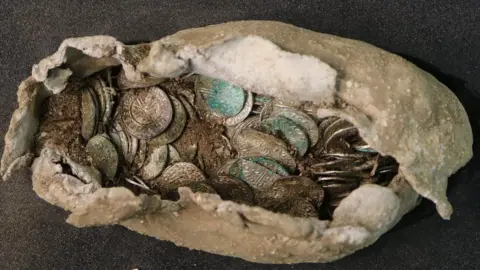 Oxford Cotswold Archaeology The cloth package containing hundreds of historic coins rests on black surface. The coin faces are detailed while some earth and dirt rest inside the package.