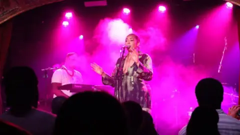Kingpin @djkingpin33 A woman bathed in purple light sings into a microphone while holding up both her hands on stage. A keyboard player can be seen to her left and a crowd of people watching in the foreground.