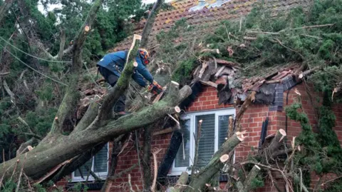 Getty A contractor wearing a hard hat uses a chainsaw to clear a tree which has fallen on to the tiled roof of a house in Burnham-on-Sea after strong winds in December 2024.