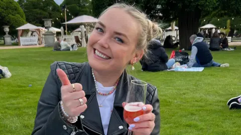 Georgie Swallow Georgie does a thumbs up to the camera, wearing a leather jacket, and holding a drink.
