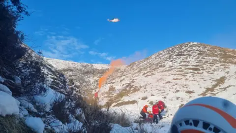 Edale MRT A helicopter approaching the scene of a rescue in a snowy mountain valley, with a smoke flare guiding the way in