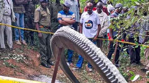 Getty Images A crowd, including police officers, stand behind yellow tape looking on at a giant metal ring which fell from space on to farmland in Mukuku, Kenya