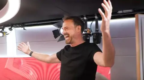 PA/ Heart Breakfast Jamie Theakston dressed in a black T-shirt and with arms outstretched arriving on the radio set of Friday's Heart Breakfast show
